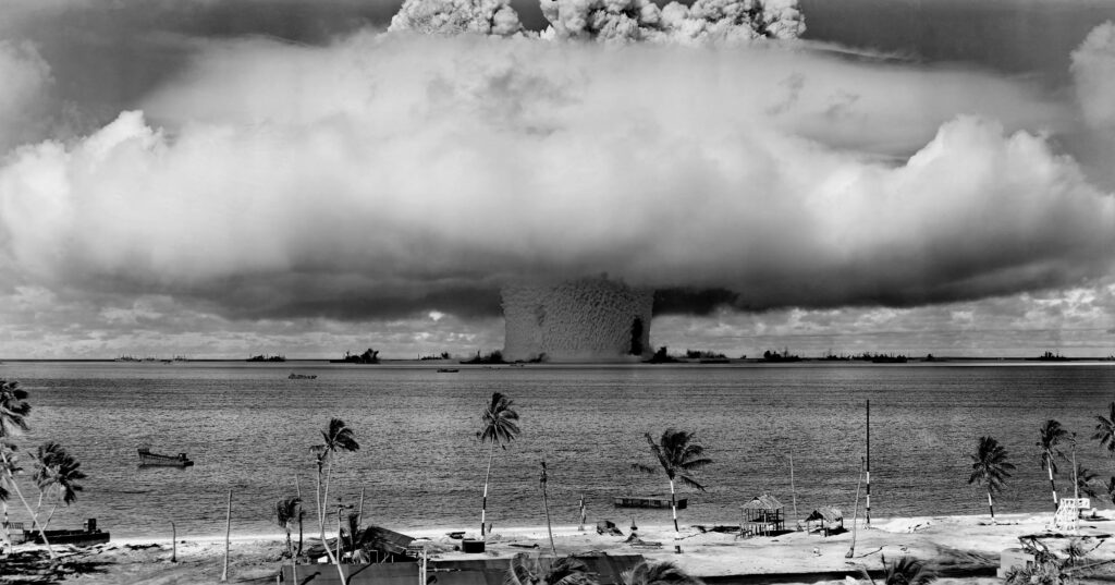 nuclear-weapons-test-nuclear-weapon-weapons-test-explosion-73909-73909 A black-and-white image of a nuclear explosion seen from a tropical beach with coconut trees.